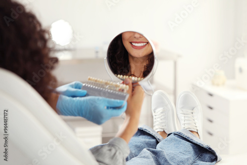 Patient looking in mirror while doctor holding teeth color chart in clinic, closeup. Dental veneers