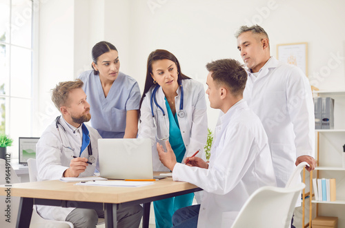 Group of professional doctors sitting with laptop at the desk on meeting discussing diagnosis, checking test results or planning treatment in clinic. Medical staff, healthcare and teamwork concept.