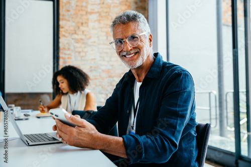 Seasoned business executive confidently working in a modern office setting, smiling while using a smartphone and laptop