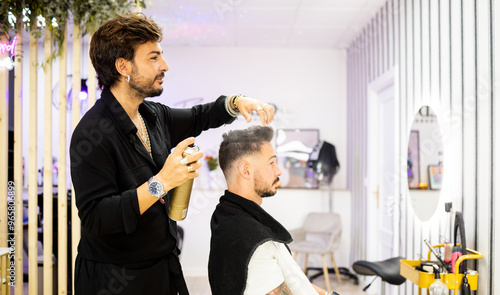 An adult hairdresser with a beard and black clothes is using a can of spray hairspray on a client's hair to set the finished hairstyle.Concept of spray hairsprays or fixatives.