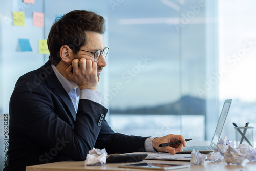 Businessman sitting at office desk appears bored while working on accounting tasks. He is using laptop and holding pen, surrounded by crumpled paper. Represents tedious office work and frustration.