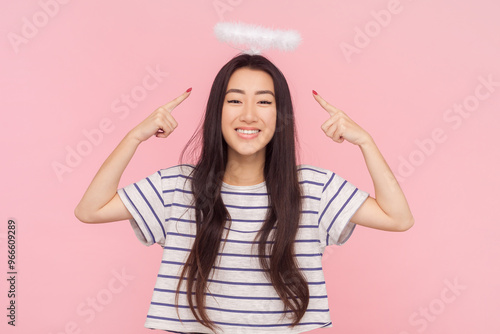 Portrait of smiling satisfied woman with long brunette hair standing pointing at her nimb expressing happiness, wearing striped T-shirt. Indoor studio shot isolated on pink background.