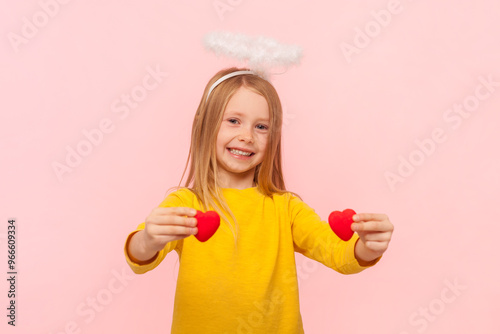 Portrait of smiling delighted blonde little girl with nimb over head showing small hearts looking at camera with happiness, wearing yellow jumper. Indoor studio shot isolated on pink background.