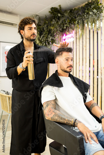 Vertical photo of an adult hairdresser with a beard and black clothes is using a can of spray hairspray on a client's hair to set the finished hairstyle. Spray lacquer or fixatives concept.