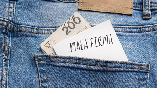 White card with a handwritten inscription "Mała Firma", put into the pocket of blue pants jeasnow, next to Polish banknotes PLN (selective focus), translation: Small Business