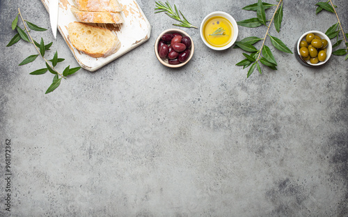 Composition with sliced fresh ciabatta on cutting board, green and brown olives, olive oil with rosemary, olive tree branches on gray concrete stone rustic background top view, copyspace border