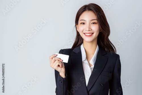 A young Asian woman smiles brightly while holding a business card, exuding confidence in her formal attire