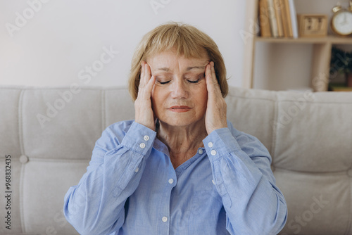 Portrait of senior woman sitting on a sofa at home with a headache, feeling pain and with an expression of being unwell