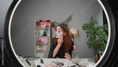 Young woman beauty blogger in witch costume at home films video tutorial for halloween party makeup on smartphone, wiping off makeup with cotton and liquid.