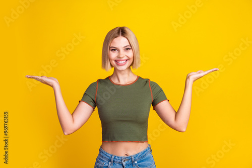 Photo of cheerful girl with bob hair dressed khaki t-shirt palms comparing products empty space isolated on yellow color background