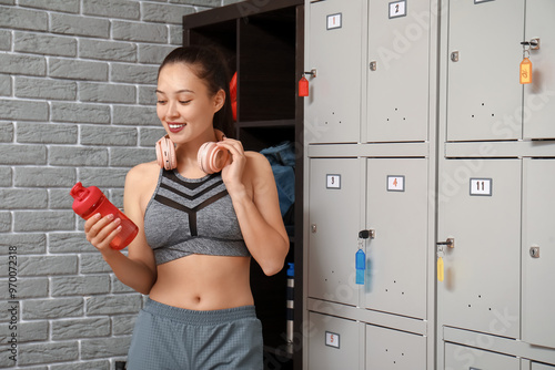 Sporty young Asian woman with water bottle and headphones in locker room