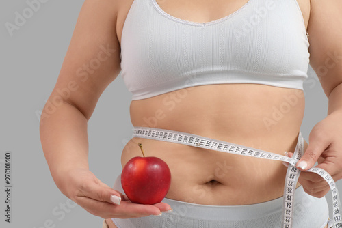 Overweight young woman with measuring tape and fresh apple on grey background, closeup. Weight gain concept