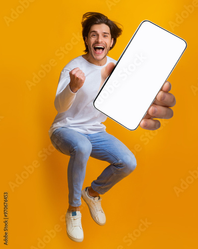 New awesome mobile app. Jumping emotional young guy showing smartphone with blank screen on orange studio background, mockup for application or website design