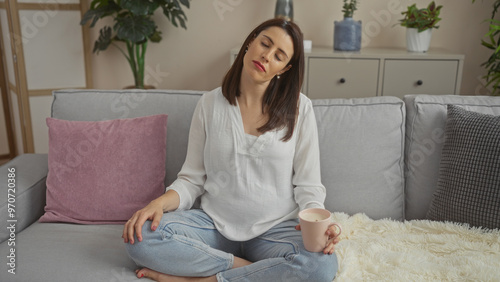 A relaxed woman sits on a cozy sofa in a modern living room, holding a cup and appearing serene and contemplative.