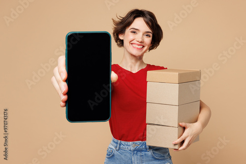 Young smiling happy woman she wears red t-shirt casual clothes hold stack cardboard boxes use blank screen mobile cell phone isolated on plain pastel light beige background studio. Lifestyle concept.