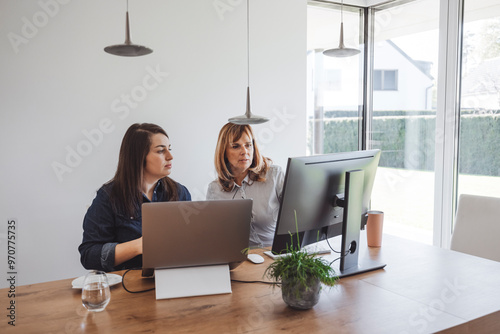 Mother and daughter collaborating at a modern workspace, one using a laptop and the other looking at a desktop monitor. The setting features a bright, airy room with large windows and minimalistic dec