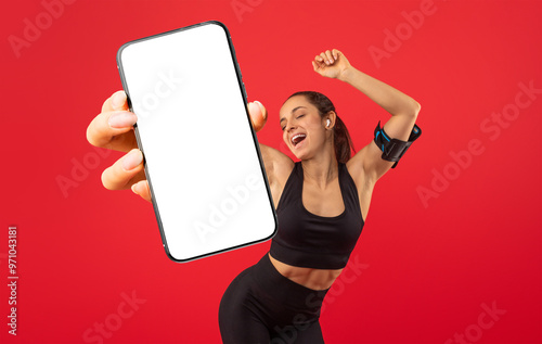 A young woman is joyfully dancing against a bright red backdrop while holding a smartphone. She wears athletic clothing and has a sporty arm band, radiating energy and positivity.