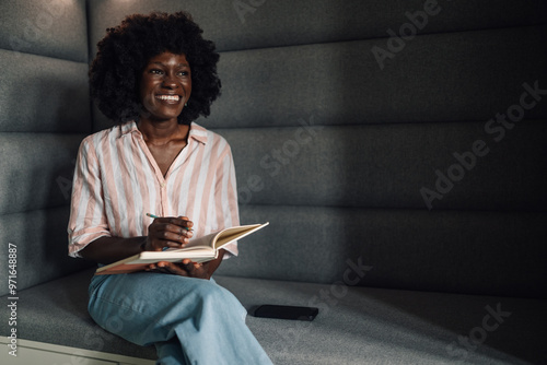 Casual diverse manager on sofa writing in textbook and looking away