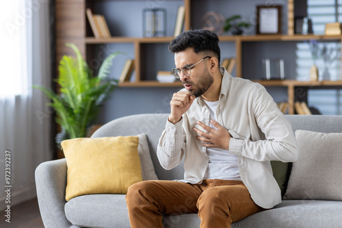 Muslim young man sitting on the sofa at home, holding his chest with his hand, feeling severe pain, coughing and covering his mouth with his hand