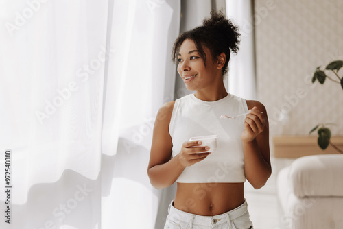 Young woman enjoying yogurt and looking out the window in a modern apartment