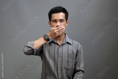 A young man covered his mouth with his hand, as if holding himself back from speaking. His facial expression showed contemplation. Isolated on gray background.