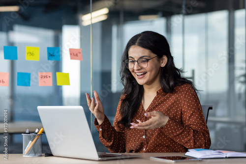 Young business woman inside office using laptop for video call, female worker smiling and talking with colleagues remotely in online meeting, joyful female worker with computer.