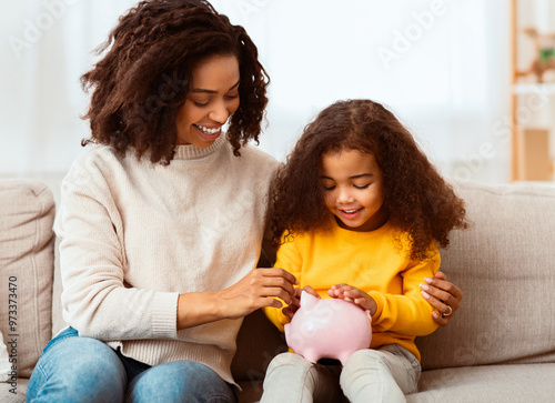 Family Savings. Cheerful Afro Mother Teaching Little Daughter How To Save Money Holding Piggybank Sitting On Sofa At Home