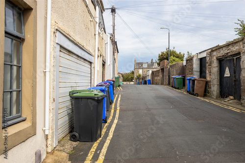 UK waste wheelie wheeled bin stored along roadside with double yellow lines and garage entrances on town side street between houses