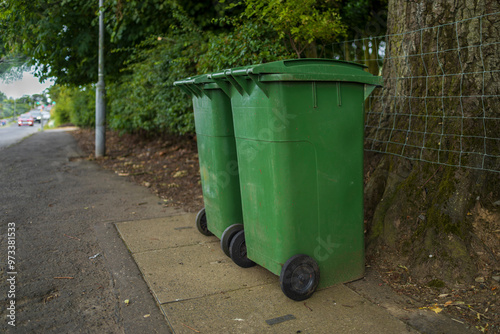 UK green waste wheelie wheeled bins on pavement beside tree trunk on busy road