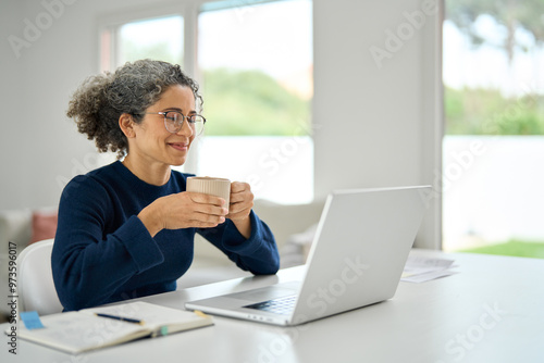 Happy mature woman sitting at home office table drinking coffee looking at laptop. Smiling relaxed middle aged older lady hybrid working on computer watching elearning webinar in modern house.