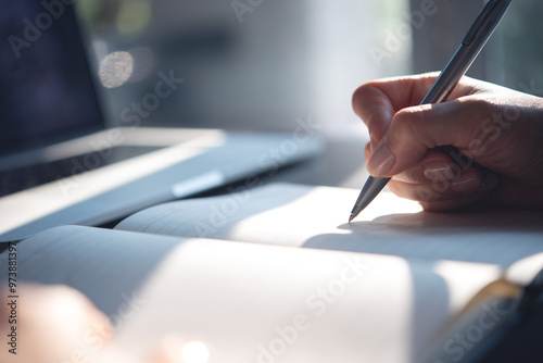 Close up of woman hand with a pen writing on notebook with laptop computer on office table. Business woman planning, making a note reminder. Student studying online, e-learning