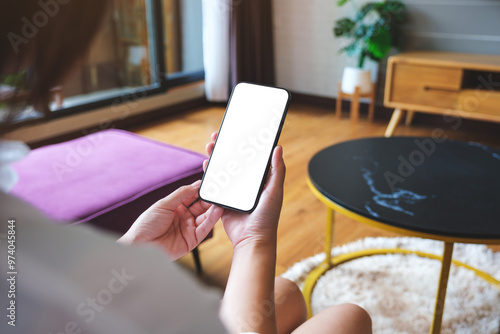 Mockup image of a woman holding mobile phone with blank desktop white screen at home