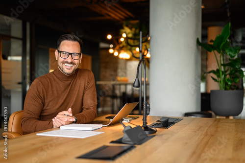 Portrait of inspired male entrepreneur in glasses working over laptop while sitting at desk in office