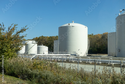 Large white storage tanks at an industrial facility surrounded by foliage under a clear blue sky, signifying robust industrial storage capacities