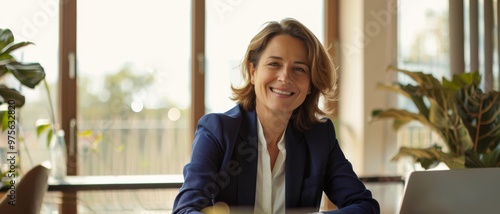 Professional woman in a suit sits confidently at her desk with a welcoming smile in a sunlit, modern office, exuding warmth and approachability.