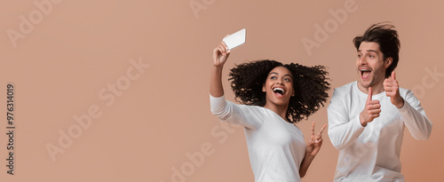 Joyful interracial couple taking selfie while jumping, showing thumbs up and peace gestures, having fun together over yellow background in studio