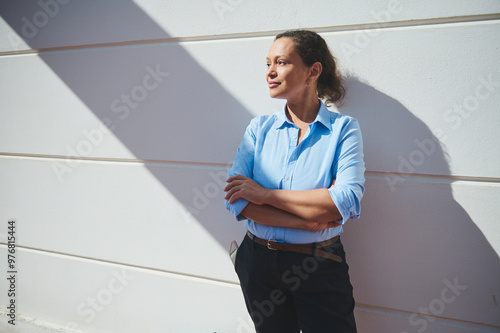 Confident woman in blue shirt posing against a white wall under sunlight. Relaxed expression and crossed arms convey empowerment and self-assurance in urban setting.