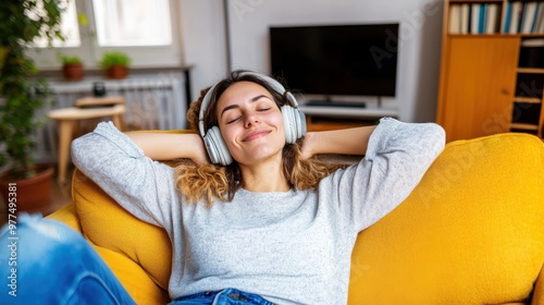 A young woman sits back on a mustard-colored sofa, eyes closed and completely immersed in music from her headphones, radiating joy and relaxation.