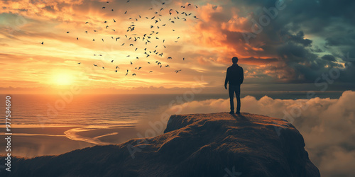 A solitary man stands on a cliff, silhouetted against a vibrant sunset sky, with a flock of birds in flight adding to the dramatic scenery.