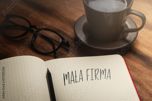 A handwritten inscription "Mała Firma" on a grille of an open notebook on a wooden countertop, next to a black pencil, a cup with coffee and glasses, a flash of light. (selective focus) 