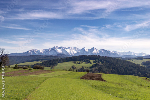 Widok na ośnieżone szczyty Tatr oraz łąki, zielone wzgórza i lasy w słoneczny dzień od strony Sromowców Wyżnych. Panorama Tatr. 