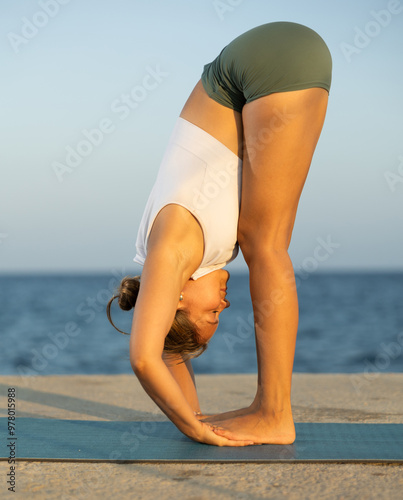 Against background of bright blue sea, slender of woman in sports clothes, hatha yoga lovers practicing oriental system of exercises. Woman performs Parivritta Uttanasana, Uttanasana