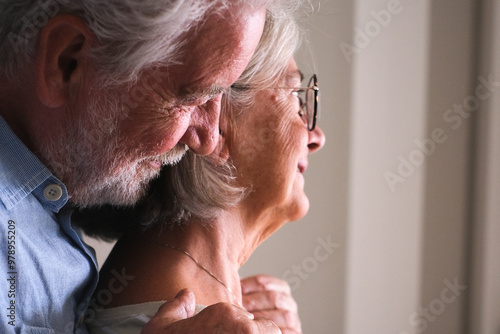 Side view of an elderly couple embracing from behind in their home, with dreamy and loving expressions. The senior man wraps his mature wife in protection and affection, showcasing their enduring love