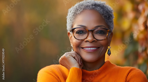 A joyful senior black woman wears stylish glasses and an orange sweater, with a warm smile that radiates happiness and confidence in a serene setting