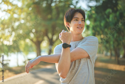 Happy sporty asian young male in sportswear stretching his arms or warming up his body before running or jogging in the green park morning.