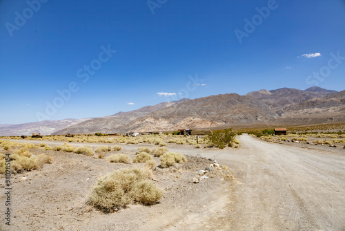 The ghost town of Ballarat, near Death Valley, California, sits abandoned in the desolate desert landscape, surrounded by rugged mountains and dry scrubland under a clear blue sky.