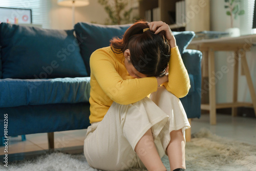 Young woman sitting on the floor in her living room, hugging her knees and burying her head, looking dejected and despondent