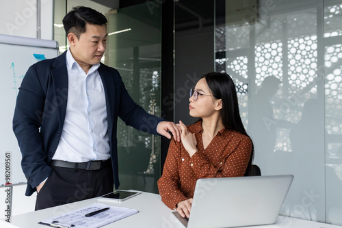 Office scene with male boss touching female subordinate's shoulder. Female employee looks uncomfortable, highlighting inappropriate workplace behavior.