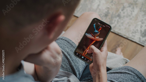 Teen boy scrolls through and watches social network feed with short videos using mobile phone while sitting on sofa. Smartphone app with various video content displayed on phone screen. Close Up.