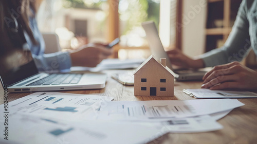 A collaborative workspace with a wooden house model on documents, laptops, and people discussing real estate or home-related matters.